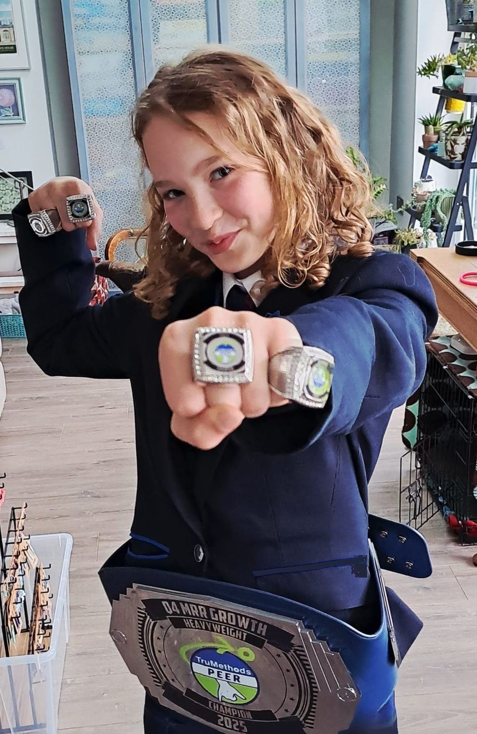 Ben’s daughter grinning at the camera, showing off two oversized championship rings and wearing a heavyweight champion’s title belt. Her school uniform has been edited to look generic.