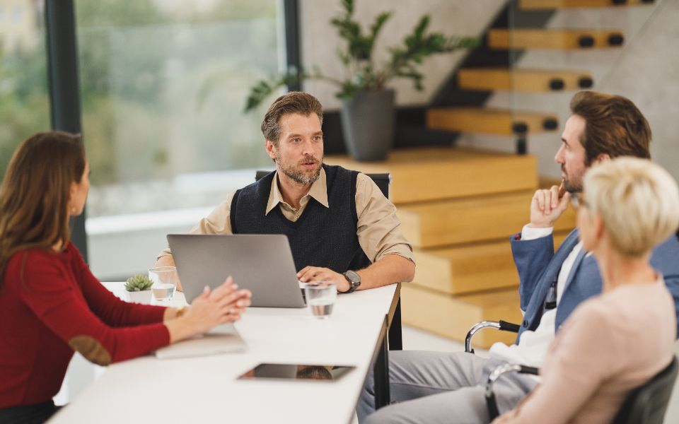 Four people in a meeting around a table, one laptop between them