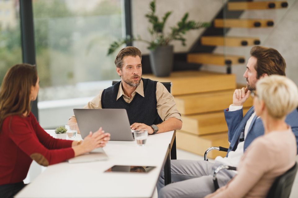 Four people in a meeting around a table, one laptop between them