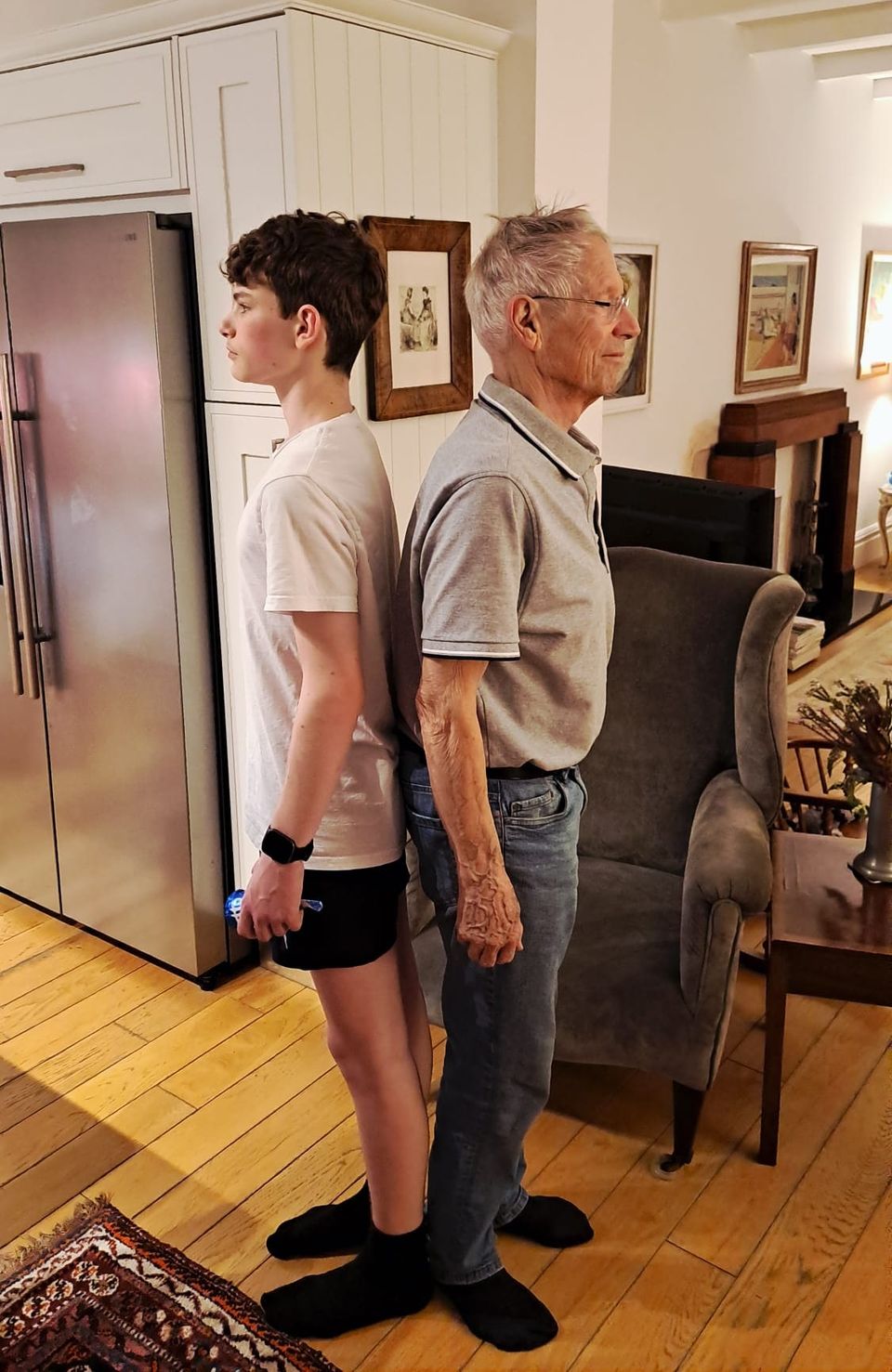 Ben’s teenage son and Ben’s dad standing back to back in a living room, comparing heights